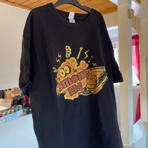Svart t-shirt med tryck - Cool svart t-shirt med ett färgglatt tryck av en tecknad bil och texten 'Smoke EM'. Perfekt för en avslappnad stil med en rolig och unik design. T-shirten har korta ärmar och en normal passform.