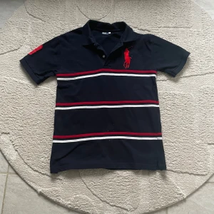 Svart pikétröja från Ralph Lauren - Säljer en stilren svart pikétröja från Ralph Lauren med röda och vita ränder. Tröjan har korta ärmar och en klassisk krage med knappar. Den ikoniska röda logotypen är broderad på bröstet. Perfekt för en avslappnad men snygg look.