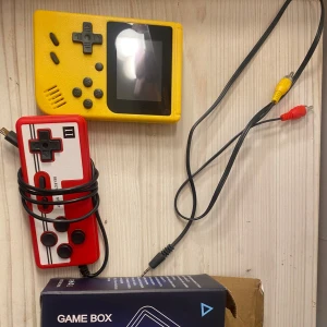 Gul bärbar spelkonsol med 400 spel - Säljer en gul bärbar spelkonsol med 400 inbyggda spel, inklusive klassiker som Super Mario och Contra. Konsolen har en svart styrkors och knappar, samt en röd och vit handkontroll. Perfekt för retrospelentusiaster! 🎮