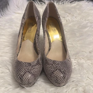 Ormskinnsmönstrade pumps från Michael Kors - Snygga pumps från Michael Kors med ormskinnsmönster i grå och beige toner. Skorna har en elegant klack och spetsig tå, vilket ger en stilren look. Perfekta för att lyfta din outfit med en touch av lyx.