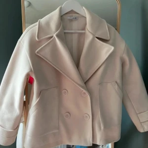 Beige Design by si kappa - Säljer en stilren beige kappa med dubbelknäppning och breda slag. Jackan har en klassisk design med långa ärmar och två fickor framtill. Perfekt för kyligare dagar! Köpt för 600 säljer billigt för vill bli av med den! 