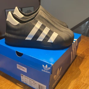 Svarta Adidas sneakers - Säljer ett par svarta Adidas sneakers med vita ränder och logotyp. Skorna har en modern design utan snörning och är tillverkade i syntetmaterial. Perfekta för en stilren look. Priset är inte hugget i sten vid snabbt köp