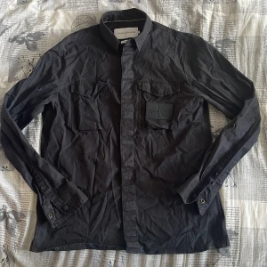 Svart skjorta från Calvin Klein - Snygg svart skjorta från Calvin Klein med långa ärmar och klassisk krage. Skjortan har två bröstfickor med lock och knappar framtill. Perfekt för en stilren look. Lite strynklig bara det ordnar sig med stryka det. 