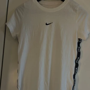 Vit t-shirt från Nike - Snygg vit t-shirt från Nike med korta ärmar och det klassiska Nike-loggan på bröstet. Sidan har en cool detalj med texten 'Nike' längs med en svart rand. 