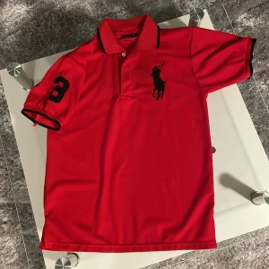 Ralph Lauren Polo Tröja - Snygg röd pikétröja från Ralph Lauren med klassisk krage och knappar. Tröjan har ett stort svart broderat logotypmotiv på framsidan och nummerdetalj på ärmen. Perfekt för en stilren look. Storleken är medium och har knappt använt den.