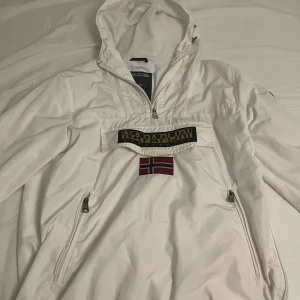 Vit anorak från Napapijri - Säljer en stilren vit anorak från Napapijri med en stor logotyp och norsk flagga på framsidan. Jackan har en praktisk huva och dragkedjor på sidorna för enkel på- och avtagning. Perfekt för kyligare dagar.