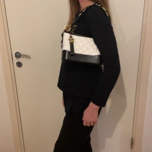 Vit och svart quiltad axelväska från Chanel - Snygg axelväska från Chanel i vit och svart med quiltad design. Väskan har en bred axelrem med guldfärgade bokstäver och detaljer. Perfekt för att ge en lyxig touch till din outfit.