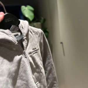 Grå hoodie från Peak Performance - Säljer en stilren grå hoodie från Peak Performance. Den har en dragkedja framtill och en praktisk huva. Perfekt för en avslappnad look. Peak Performance-loggan är diskret placerad på bröstet.