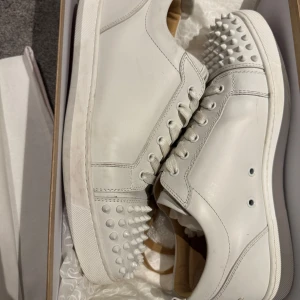 Vita sneakers med nitar från Christian Louboutin - Snygga vita sneakers från Christian Louboutin med ikoniska röda sulor och coola nitar på sidorna och framtill. Skorna har snörning och är tillverkade i skinn, vilket ger en lyxig känsla. Perfekta för dig som vill sticka ut med stil. PRISET EJ HUGGET I STEN!!!