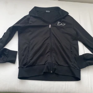 Svart jacka från Emporio Armani - Snygg svart jacka från Emporio Armani EA7 med dragkedja framtill. Jackan har en stilren design med logotyp på bröstet och ribbade muddar vid ärmslut och nederkant. Perfekt för en casual look.