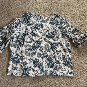 Mönstrad blus från H&M - Säljer en mönstrad blus från H&M i vitt och blått med paisley- och blommönster. Blusen har trekvartsärmar och en lös passform i viskos. Perfekt för en avslappnad stil.
