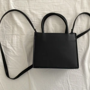 Svart axelväska - Snygg och stilren svart axelväska i syntetmaterial. Väskan har en enkel design med långa axelremmar, perfekt för en minimalistisk look. Passar till många olika outfits och tillfällen.