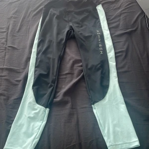 Svarta och mintgröna leggings från Röhnisch - Snygga leggings från Röhnisch i svart med mintgröna paneler längs sidorna. Perfekta för träning med en tight passform och hög midja. Tillverkade i ett stretchigt syntetmaterial för optimal rörelsefrihet.