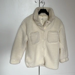 Vit teddyjacka från H&M - Mysig vit teddyjacka från H&M med två stora bröstfickor och knäppning framtill. Perfekt för kyliga dagar med sin fluffiga och varma design. Jackan har långa ärmar och en bekväm passform. Skulle säga att jackan passar Xs. Aldrig använd.