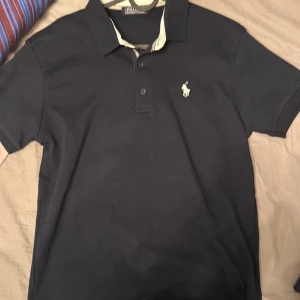 Svart pikétröja från Polo Ralph Lauren - Snygg svart pikétröja från Polo Ralph Lauren med klassisk krage och knappar framtill. Tröjan har det ikoniska broderade logotypen på bröstet. Perfekt för en stilren look.