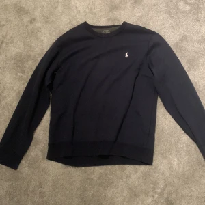 Mörkblå tröja från Ralph Lauren - Säljer en stilren mörkblå tröja från Ralph Lauren med det klassiska logotypbroderiet på bröstet. Tröjan har långa ärmar och en rund halsringning, perfekt för en avslappnad men ändå elegant look. Passar L 