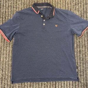 Blå pikétröja från Jack & Jones - Snygg blå pikétröja från Jack & Jones med röda och vita detaljer på krage och ärmslut. Tröjan har en klassisk pikédesign med knappar framtill och en liten broderad logotyp på bröstet.