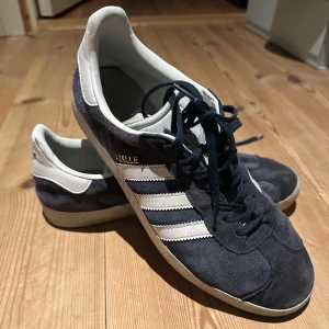 Adidas Gazelle i mörkblå mocka 42 2/3 - Säljer ett par klassiska Adidas Gazelle i mörkblå mocka med vita detaljer och snörning. Skorna har den ikoniska tre-randiga designen på sidan och en bekväm passform. Ganska slitna, därav priset.