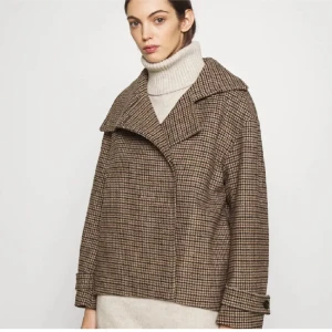 Rutig kappa med bred krage - Snygg rutig kappa i bruna och beige toner med bred krage och knappar framtill. Perfekt för höst och vår, med en klassisk och tidlös design. Passar bra till både jeans och klänning.