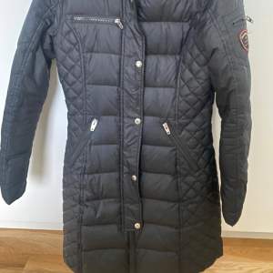 Snygg svart quiltad jacka från Rockandblue med dragkedja och knappar. Jackan har en huva och flera praktiska fickor med dragkedjor. Perfekt för kyligare dagar.