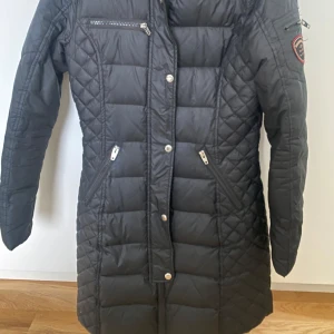 Svart quiltad jacka från Rockandblue - Snygg svart quiltad jacka från Rockandblue med dragkedja och knappar. Jackan har en huva och flera praktiska fickor med dragkedjor. Perfekt för kyligare dagar.