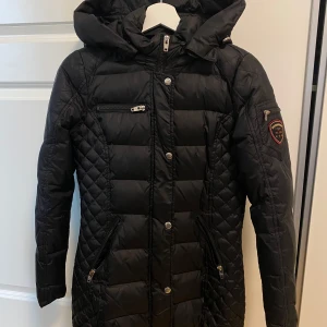 Svart pufferjacka från Rockandblue - Snygg svart pufferjacka från Rockandblue med huva och dragkedja. Jackan har quiltade detaljer och flera praktiska fickor med dragkedjor. Perfekt för kyliga dagar.