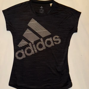 Svart tränings-tshirt från Adidas - Snygg svart t-shirt från Adidas med stort vitt logotryck på framsidan. Perfekt för träning eller en avslappnad stil. Tillverkad i ett mjukt och bekvämt material.