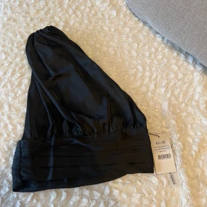 Svart oneshoulder topp - Snygg svart oneshoulder topp i ett glansigt material. Perfekt för en stilren look med en elegant touch. Toppen har en bred kant nedtill som ger en fin passform.