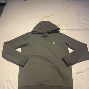 Svart hoodie från Lyle & Scott - Säljer en stilren svart hoodie från Lyle & Scott med deras ikoniska gula logga på bröstet. Tröjan har en klassisk känguruficka och en bekväm huva. Perfekt för en avslappnad look.