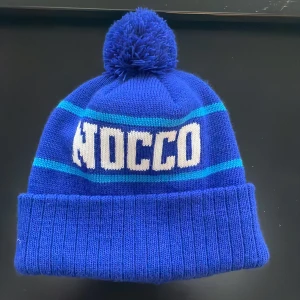 Mörkblå mössa med tofs från NOCCO - Snygg blå mössa med ribbad kant och tofs. Mössan har ljusblå ränder samt NOCCO-logga framtill. Perfekt för kalla dagar.