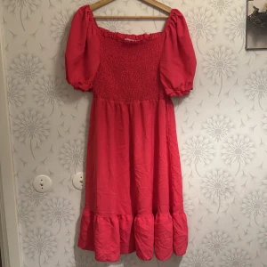 Rosa klänning med puffärmar - Charmig rosa klänning med puffiga kortärmar och smockdetaljer upptill. Klänningen har en volangkant nedtill som ger en lekfull touch. Perfekt för somriga dagar.