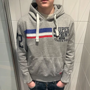 Grå hoodie från Superdry - Säljer denna grå hoodie från Superdry med tryck på bröstet, luvan och höger arm. Riktigt snygg! skick 8/10! han på bilden är 183 cm 70 kg! inga defekter!