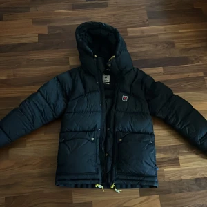 Fjällräven jacka  - Säljer en svart Fjällräven Expedition Down Lite Jacket från 1974-serien. Jackan är perfekt för kalla vinterdagar med sin tjocka dunfyllning och har en praktisk huva. Den har en dragkedja framtill och flera fickor för förvaring.