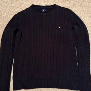 GANT tröja - Säljer en stilren svart kabelstickad tröja från GANT i premium bomull. Tröjan har en klassisk rund hals och långa ärmar. Diskret GANT-logga på bröstet.