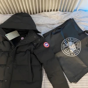 Svart dunjacka från Canada Goose - Säljer en svart dunjacka från Canada Goose med huva och knappar. Jackan har en klassisk design med varumärkets logga på ärmen. Perfekt för kalla vinterdagar.