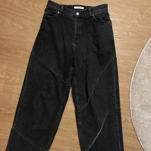 Svarta jeansbyxor från JieDa - Snygga svarta jeansbyxor från JieDa med unik sömdesign. Byxorna har en loose passform och är midwaist. De har knappar och dragkedja framtill samt två bakfickor. Perfekta för en avslappnad stil. Heelbite på högra benet och lite slitage på vänster ben