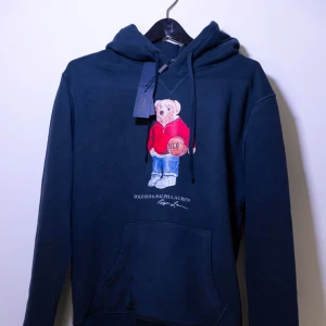 Polo Ralph Lauren Hoodie - Helt ny Polo Ralph Lauren Hoodie.Tröjan har en nallebjörn på framsidan och en klassisk känguruficka och justerbar huva. Perfekt för en avslappnad stil.