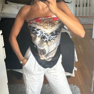 Mönstrad axelbandslös topp - Snygg axelbandslös topp med ett färgglatt mönster i rött, svart, guld och silver. Toppen har en unik design med ett scarf-liknande utseende och passar perfekt för en trendig look.