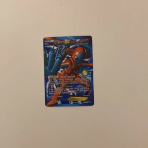 Deoxys EX Pokémonkort - Säljer ett Deoxys EX Pokémonkort med en färgglad design. Kortet har en blå och röd färgpalett med en bild av Deoxys i en dynamisk pose. Perfekt för samlare eller Pokémon-entusiaster!