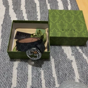 GG bälte  - Snyggt svart bälte från Gucci med det klassiska GG-mönstret. Bältet har ett stort, runt spänne i silverfärg med Gucci-logotypen. Kommer i en elegant grön låda med mönster.