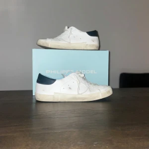 Vita sneakers från Philippe Model - Snygga vita sneakers från Philippe Model med svarta detaljer på hälen. Skorna har en klassisk design med snörning och en bekväm sula. Perfekta för en stilren look.