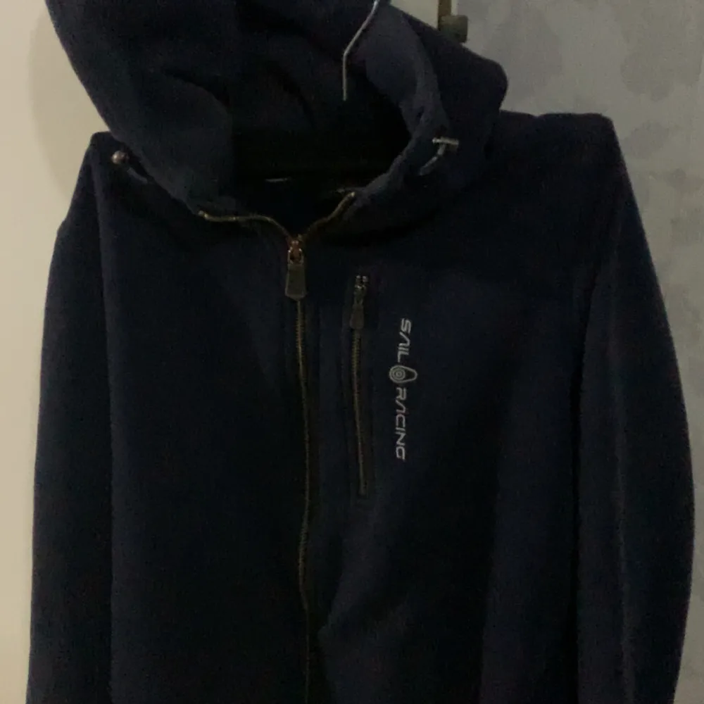 Säljer en mörkblå fleecejacka från Sail Racing med dragkedja och huva. Jackan har en broderad logotyp på bröstet och en praktisk bröstficka med dragkedja. Perfekt för kyliga dagar! Problemet är att vid händerna är lite söndriga . Takit.