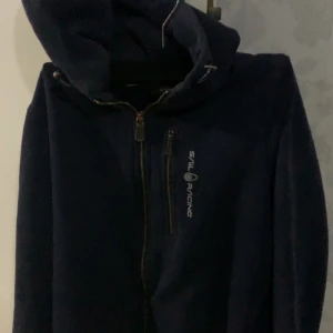 Mörkblå fleecejacka från Sail Racing - Säljer en mörkblå fleecejacka från Sail Racing med dragkedja och huva. Jackan har en broderad logotyp på bröstet och en praktisk bröstficka med dragkedja. Perfekt för kyliga dagar! Problemet är att vid händerna är lite söndriga 
