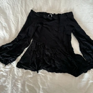 Svart offshoulder blus - Snygg svart offshoulder blus med volangdetaljer och långa ärmar. Perfekt för en trendig look.