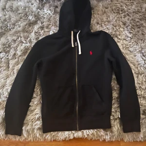 Svart hoodie med dragkedja från Ralph Lauren - Snygg svart hoodie från Ralph Lauren med dragkedja och huva. Den har en liten röd logotyp på bröstet och vita snören vid huvan. Perfekt för en avslappnad stil.