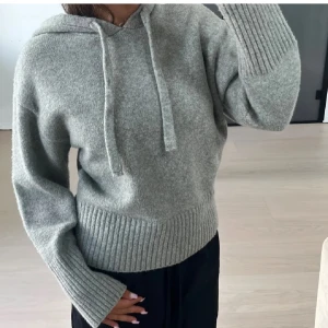Grå stickad hoodie - Mysig grå stickad hoodie med ribbade muddar och dragsko i huvan. Perfekt för kyliga dagar och avslappnade stunder. Enkelt att matcha med olika outfits.