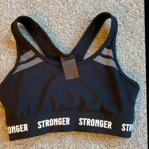 Svart sporttopp från Stronger - Snygg svart sporttopp från Stronger med vita detaljer och text. Toppen har en bekväm passform och breda axelband för extra stöd. Perfekt för träning och aktiva dagar.