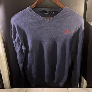Blå tröja från Polo Ralph Lauren - Säljer en stilren blå tröja från Polo Ralph Lauren i mjuk pima bomull. Tröjan har en v-ringad hals och det ikoniska röda logotypbroderiet på bröstet. Perfekt för en klassisk och avslappnad look. Hör av er vid minsta fundering🙌