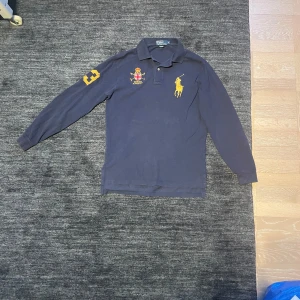 Mörkblå långärmad pikétröja från Ralph Lauren - Säljer en snygg mörkblå pikétröja från Ralph Lauren med broderad logga och emblem på bröstet. Tröjan har långa ärmar och en stor gul siffra 3 på ryggen.  I gott skick 👍