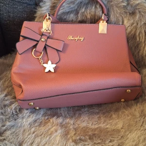 handväska med rosett och stjärndetalj - Snygg handväska i syntetmaterial med en dekorativ rosett och en charmig stjärndetalj. Väskan har guldfärgade detaljer, Perfekt för att hålla dina saker organiserade med flera fack inuti. 